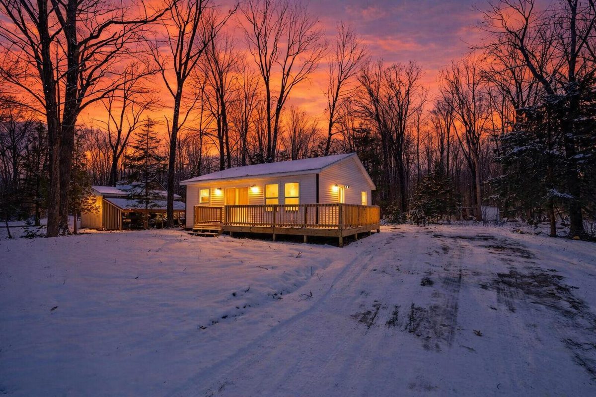 Small home with glowing windows in the snow at sunset among bare trees